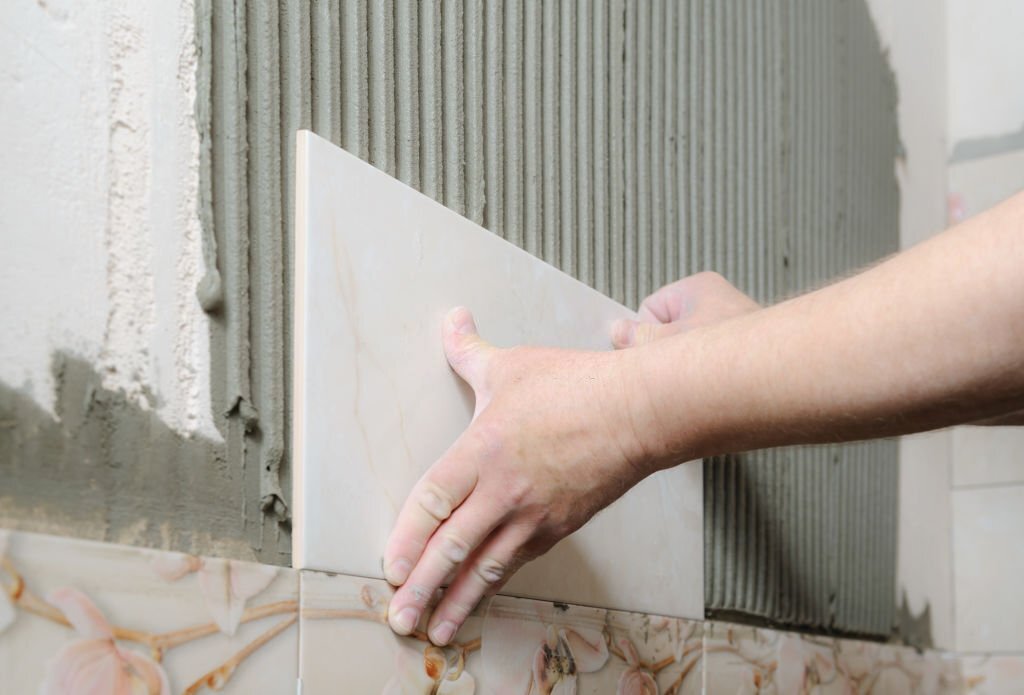 Tilers hands are  installing a ceramic tile on a wall in a bathroom.